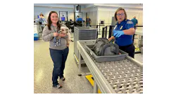 A traveler removes their pet dog from its travel carrier at the security checkpoint and sends the empty carrier into the X-ray machine as the TSA officer guides the passenger through the process.