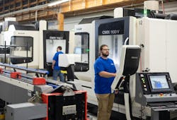 Two employees in blue shirts work on an assembly line with large machinery around them. Two employees in blue shirts work on an assembly line with large machinery around them.