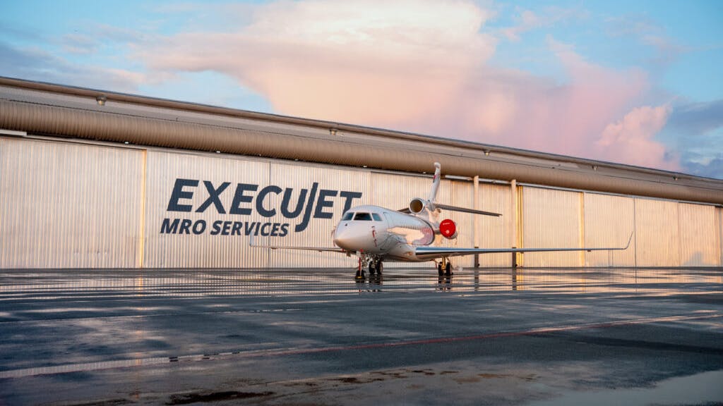 A white airplane sits on in front of a beige hangar that has a sign reading, 'ExecuJet MRO Services'