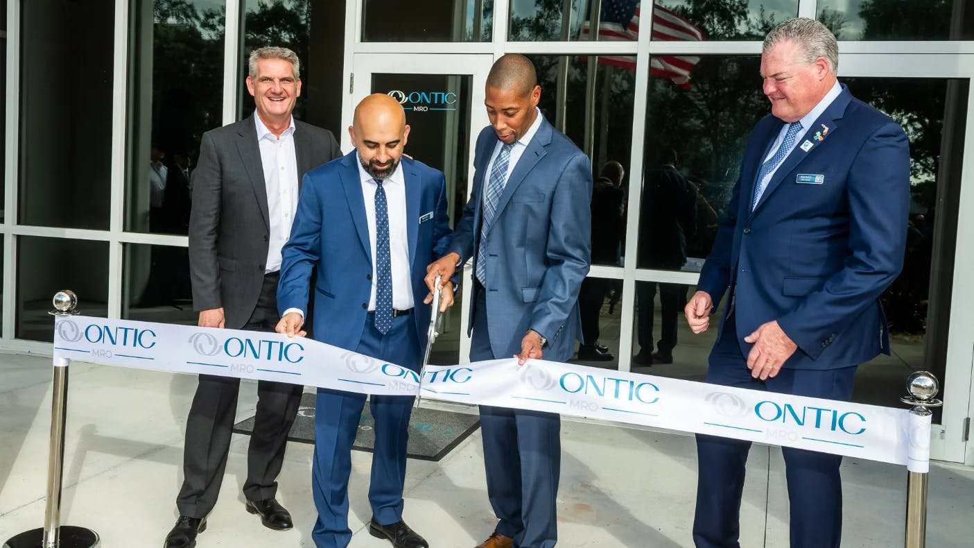 Four men wearing suits stand at a ribbon cutting ceremony with one cutting a ribbon with metal scissors