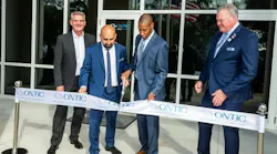 Four men wearing suits stand at a ribbon cutting ceremony with one cutting a ribbon with metal scissors