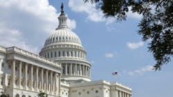 The U.S. Capitol Building with white domes and pillars and an American flag against a blue sky The U.S. Capitol Building with white domes and pillars and an American flag against a blue sky