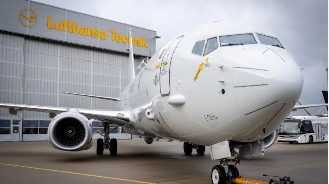 A white airplane sits in front of a grey building with yellow letters that read, 'Lufthansa Technik'