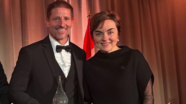 A woman in a black dress stands next to a man wearing a black suit and holding a crystal award, both smiling
