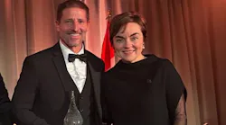 A woman in a black dress stands next to a man wearing a black suit and holding a crystal award, both smiling