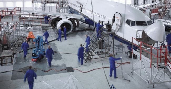 A group of maintenance technicians in blue clothing work on an airplane in an aircraft hangar