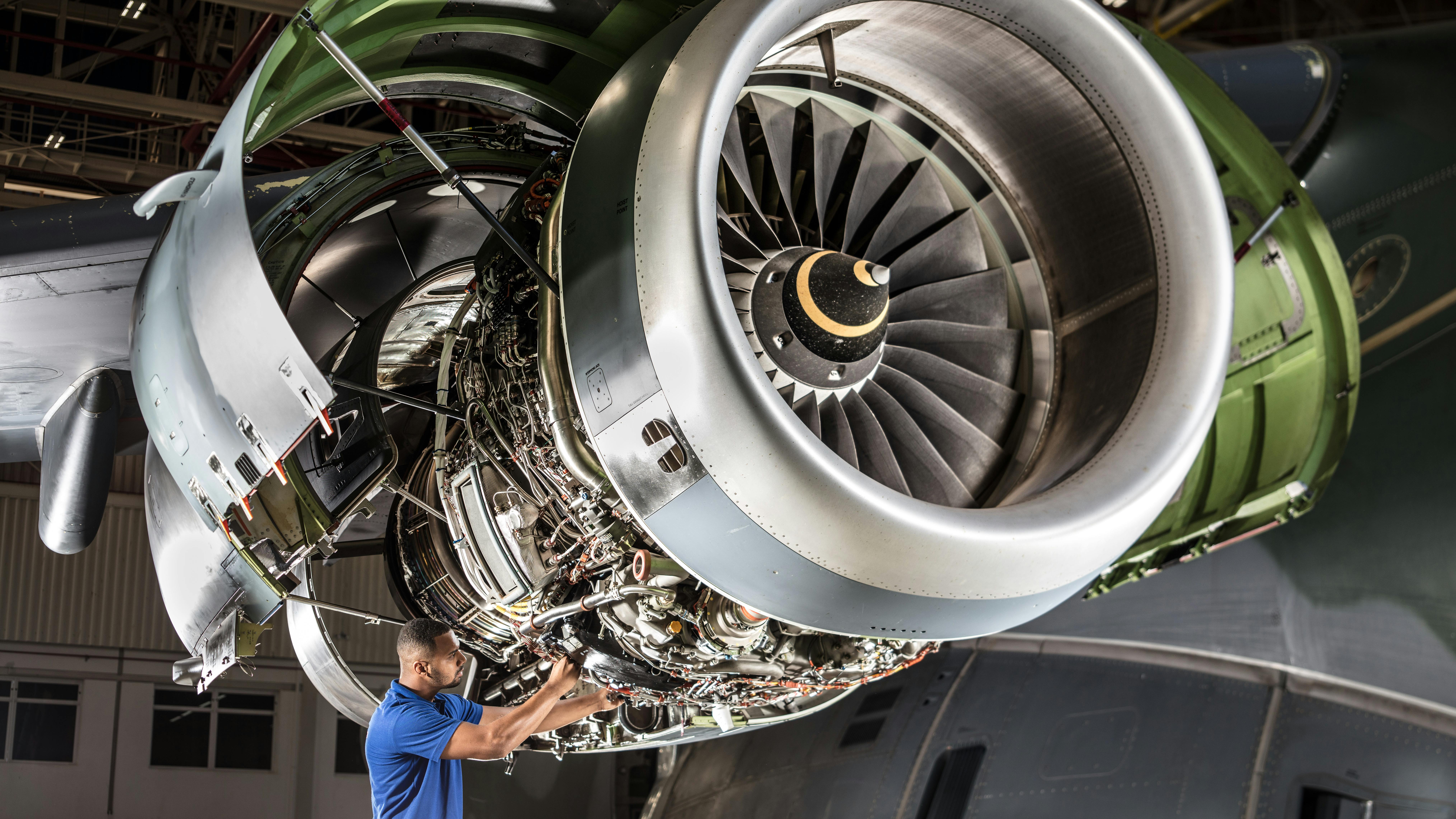 An aircraft maintenance technician in a blue shirt works on a large aircraft engine