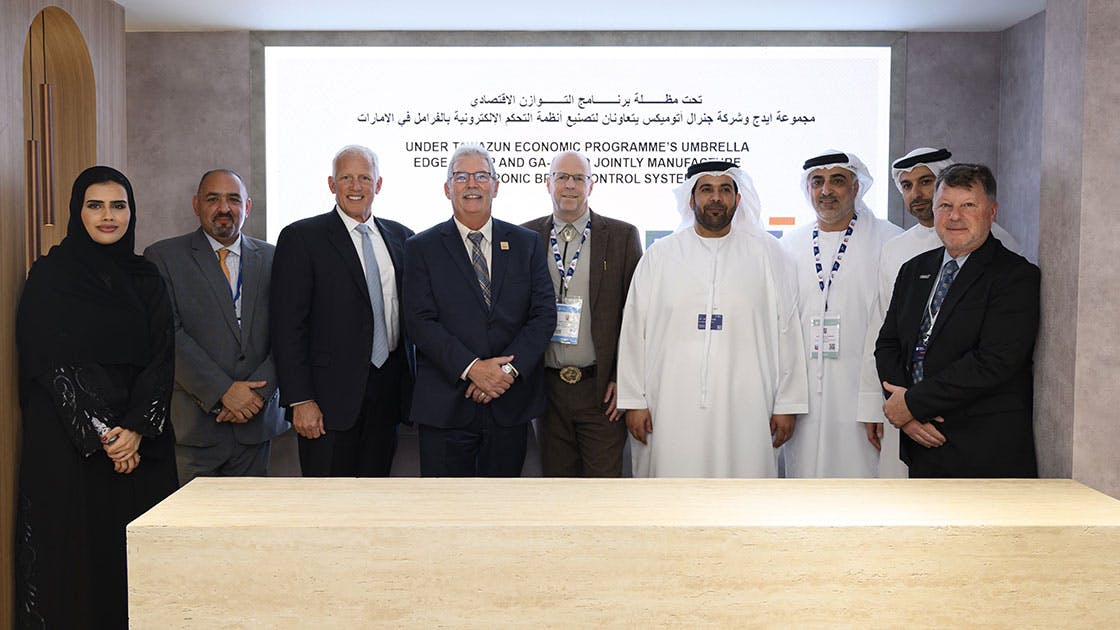 A group of people in professional attire gather around a wooden table in front of a projector screen