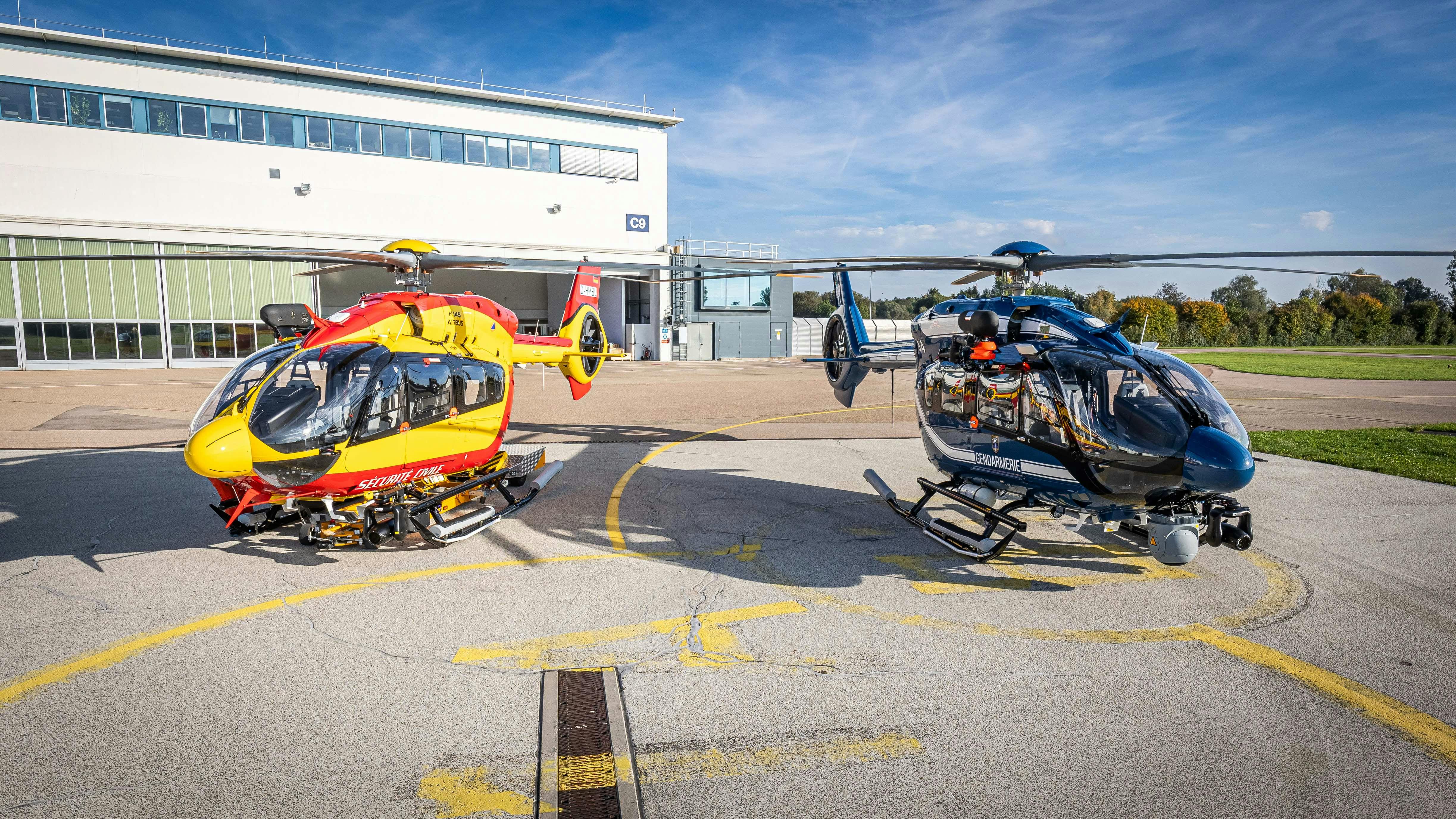 A yellow helicopter and a black helicopter sit in front of a white building