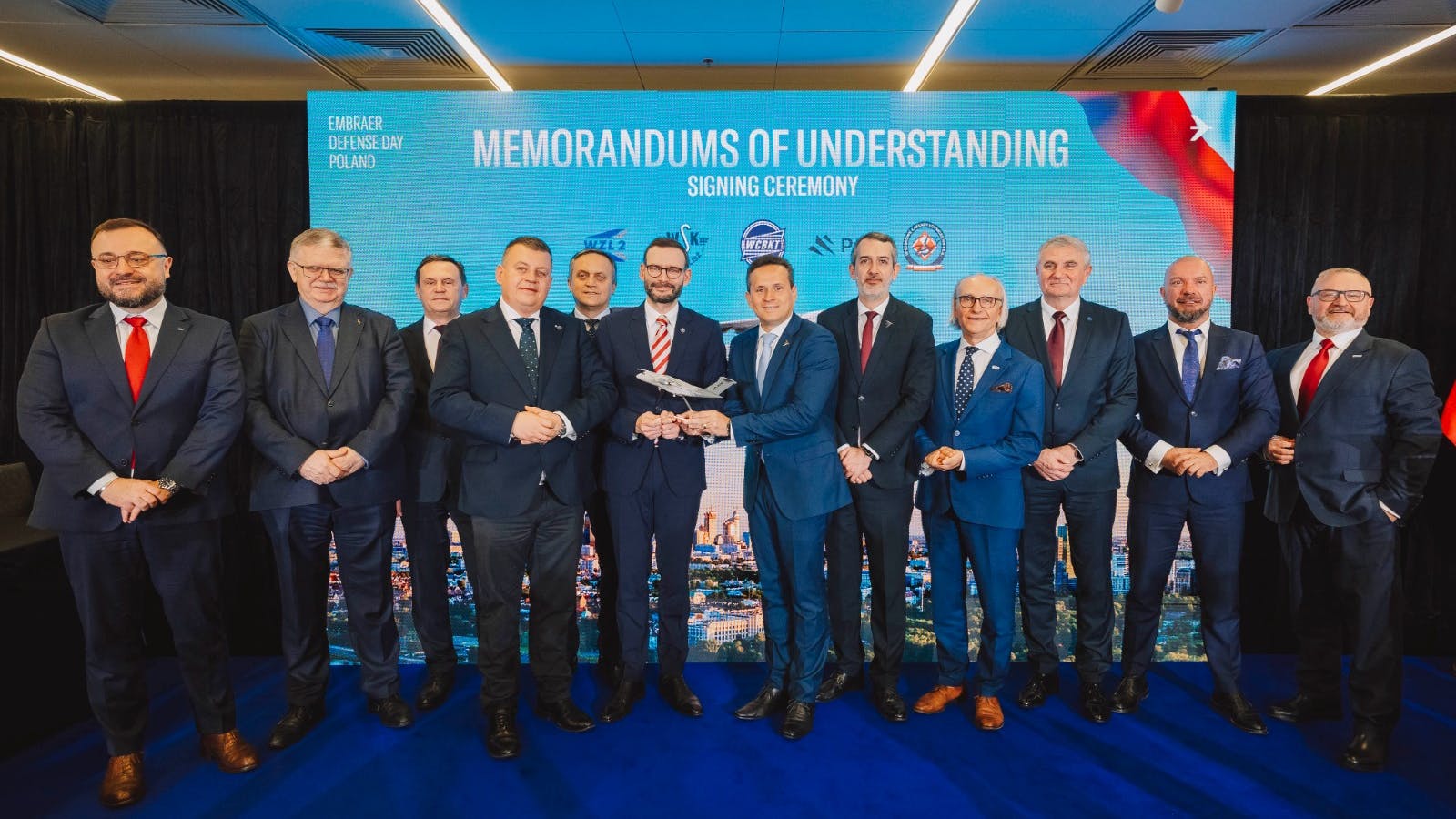 A group of professionals in business attire stand smiling in front of a banner that reads 'Memorandums of Understanding Signing Ceremony'