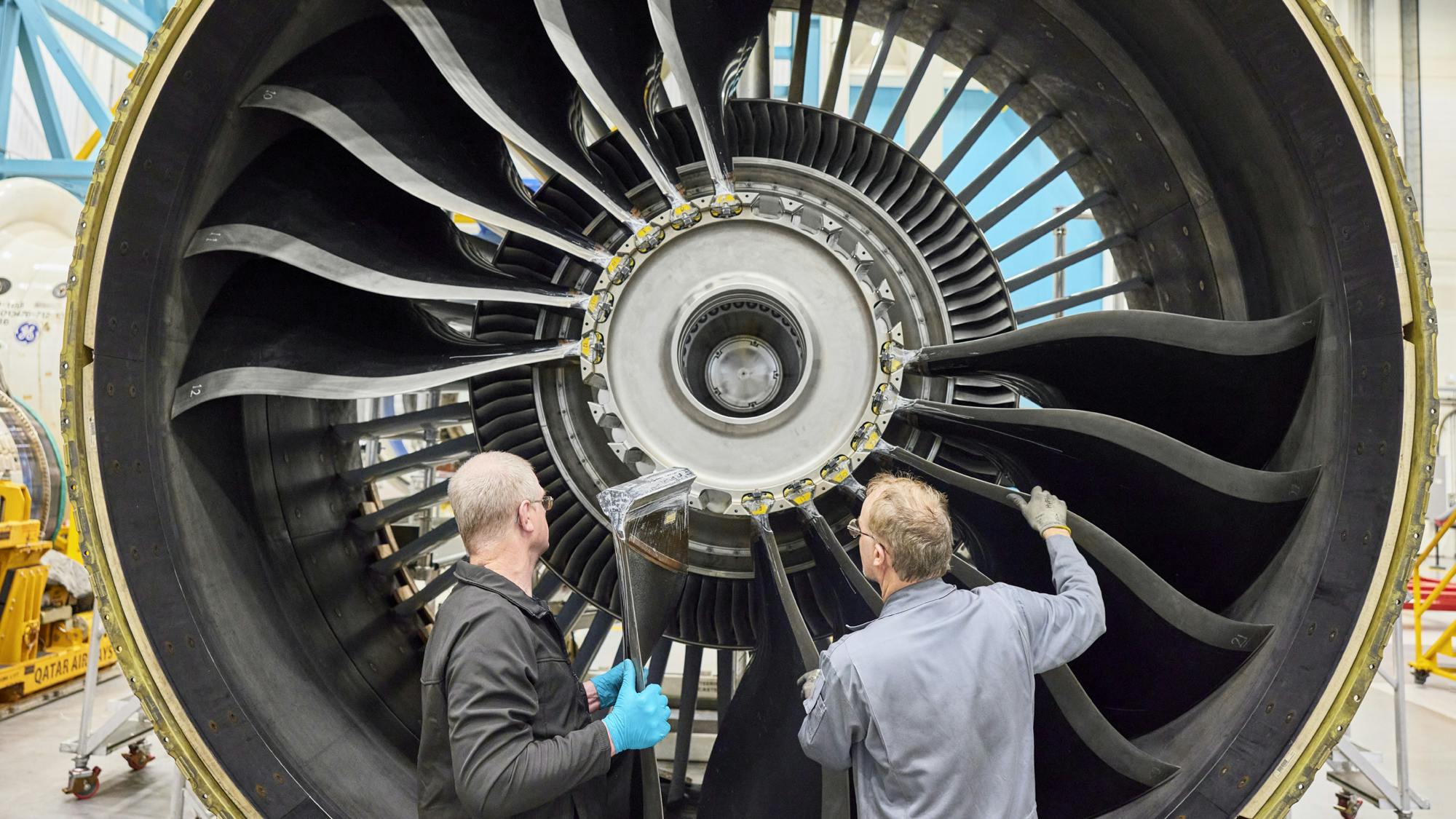 Two technicians, one wearing blue gloves, stand in front of a large aircraft engine and hold a new blade