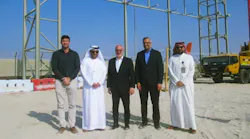 Five men in professional attire stand smiling at the camera on a construction site, with metal framework of a building behind them