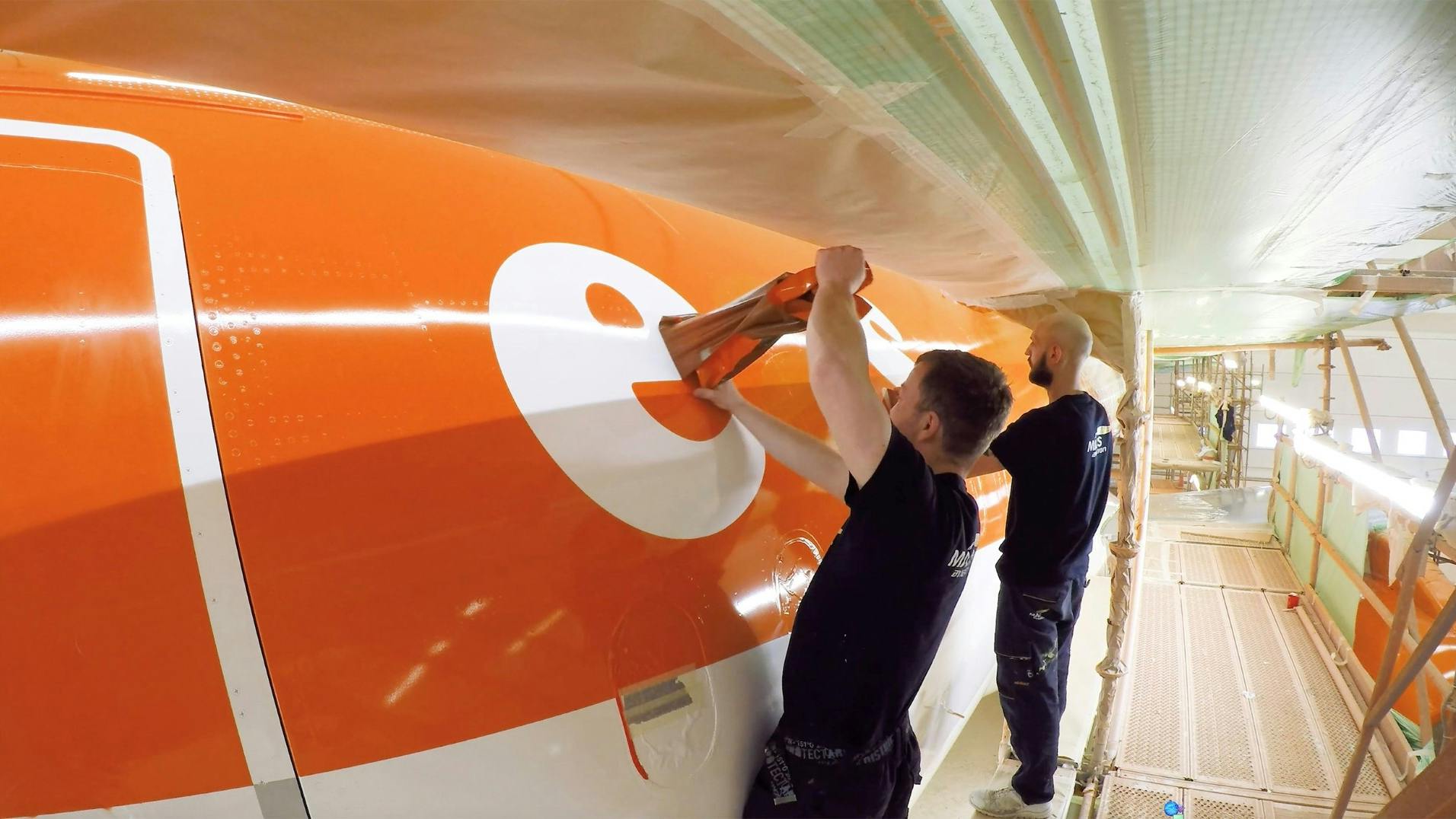 Two technicians wearing black clothing painting an orange and white airplane