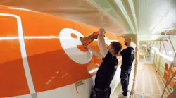 Two technicians wearing black clothing painting an orange and white airplane