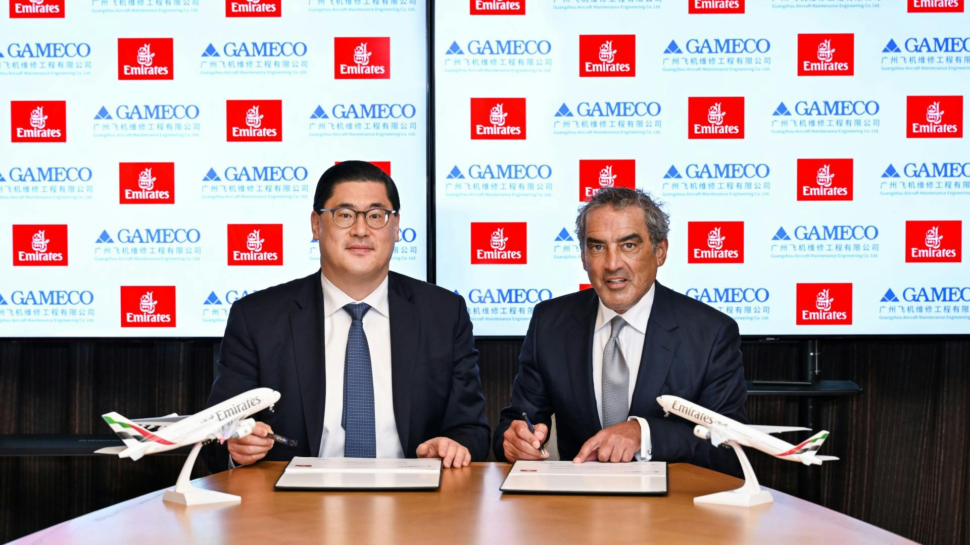 Two men wearing suits sit at a wooden table signing documents, with two airplane models on the table.