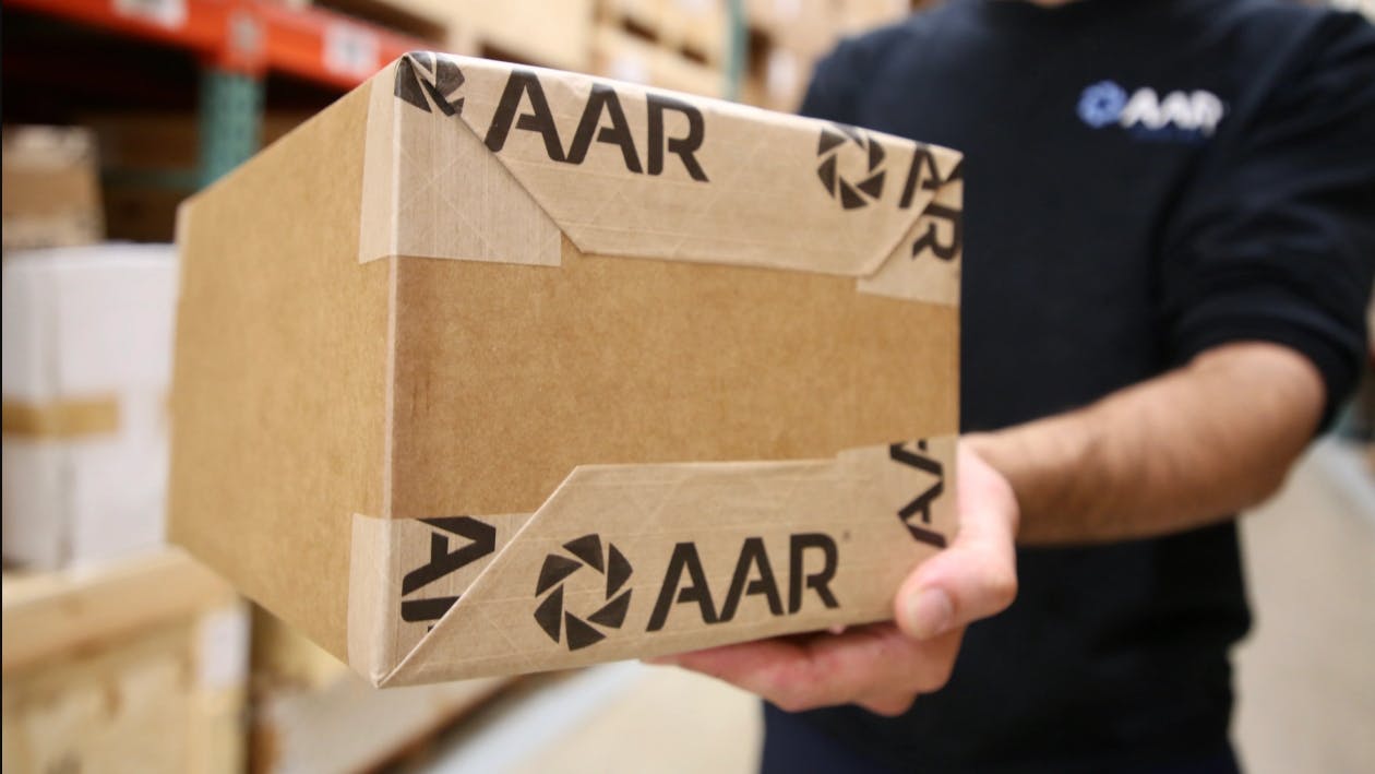 A person in a black shirt that says 'AAR' on it holding a brown shipping box with 'AAR' written on it in the same font