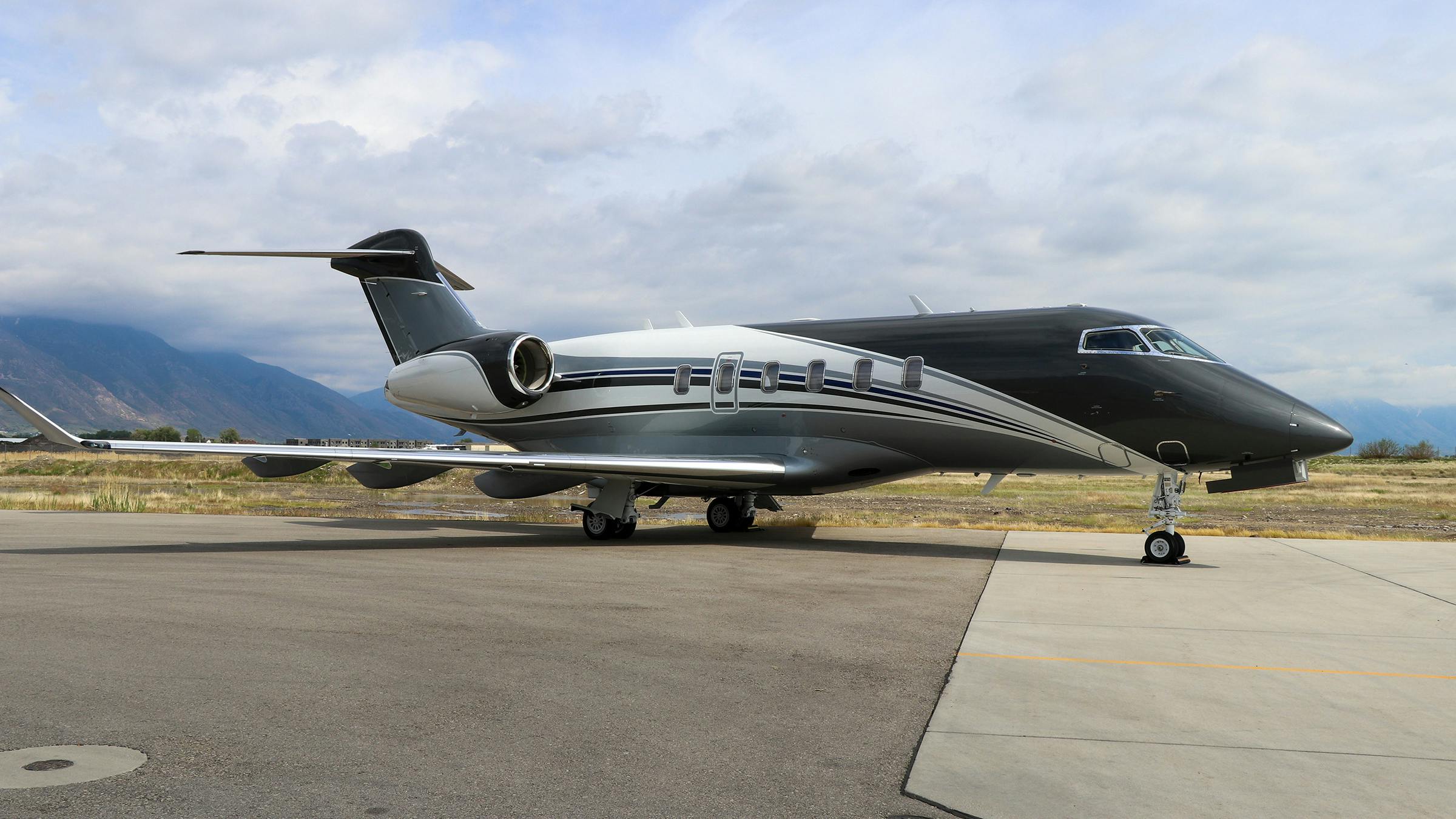 A black and white airplane with ornate stripe designs on the side sits on a tarmac with mountains and a blue sky behind it