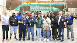 A group of employees stand in an aircraft hangar in front of a deconstructed airplane, giving thumbs-ups and smiling at the camera