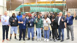 A group of employees stand in an aircraft hangar in front of a deconstructed airplane, giving thumbs-ups and smiling at the camera A group of employees stand in an aircraft hangar in front of a deconstructed airplane, giving thumbs-ups and smiling at the camera
