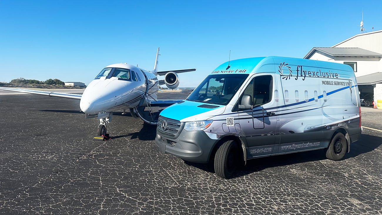 A chrome-colored van with text on it that reads 'flyExclusive' is parked next to a white jet plane on a tarmac