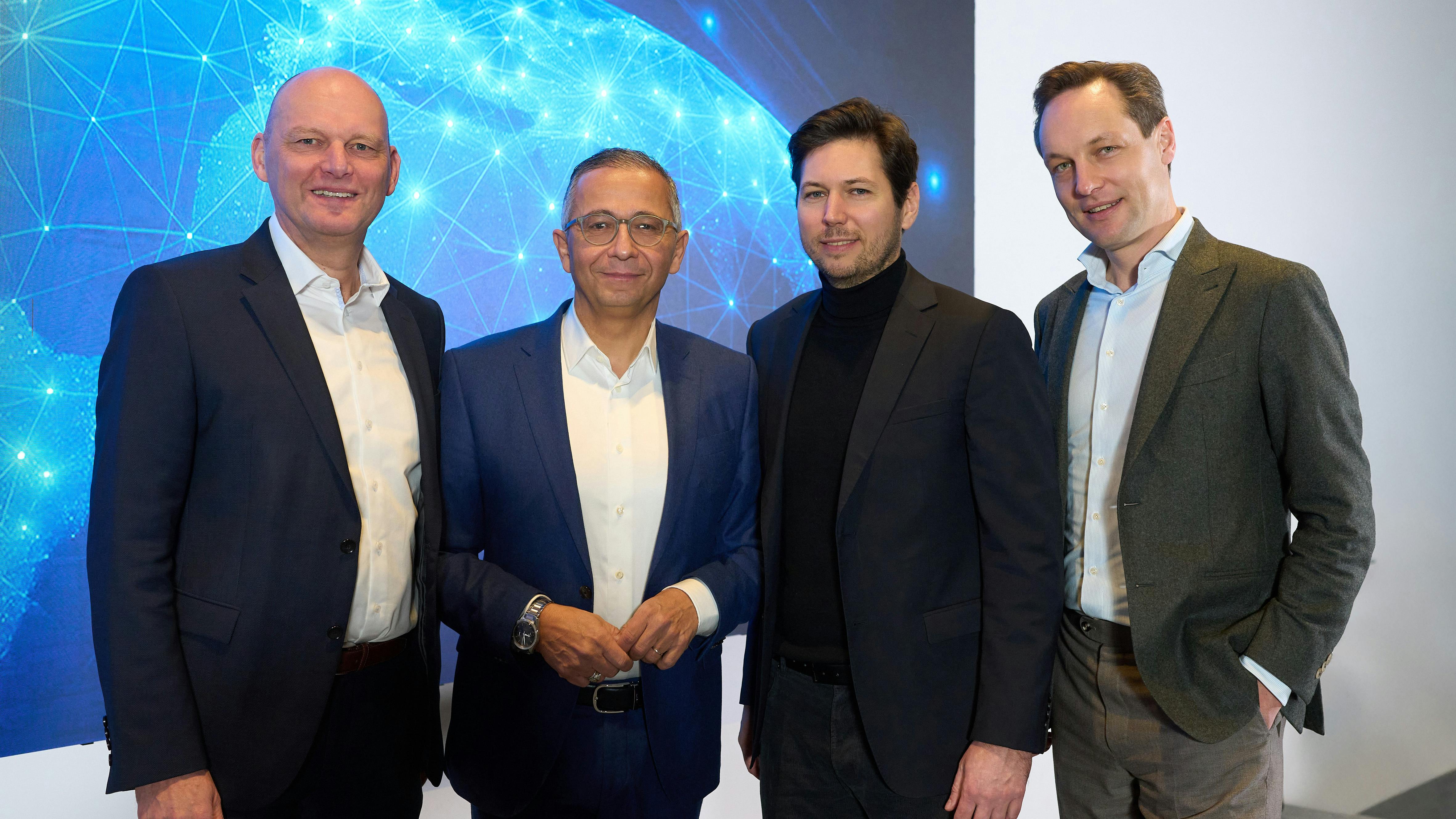Four men in business attire smile at the camera, standing in front of a bright blue design on the wall