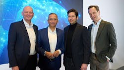 Four men in business attire smile at the camera, standing in front of a bright blue design on the wall Four men in business attire smile at the camera, standing in front of a bright blue design on the wall