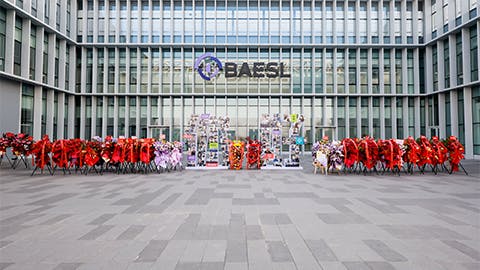 A large group of people stands outside a glass facility with the word 'BAESL' on it