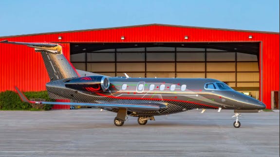A dark gray aircraft with blue and red stripes sits on a tarmac in front of a red building