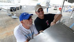 Two men work together on an aircraft's wing with another airplane behind them Two men work together on an aircraft's wing with another airplane behind them