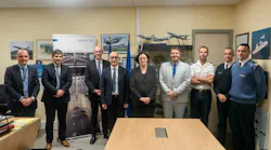 A group of people in business attire and military clothing stand behind a table in a meeting room decorated with pictures and models of airplanes