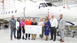 A group of people in business attire smile at the camera while holding a giant check and standing in front of a white airplane A group of people in business attire smile at the camera while holding a giant check and standing in front of a white airplane
