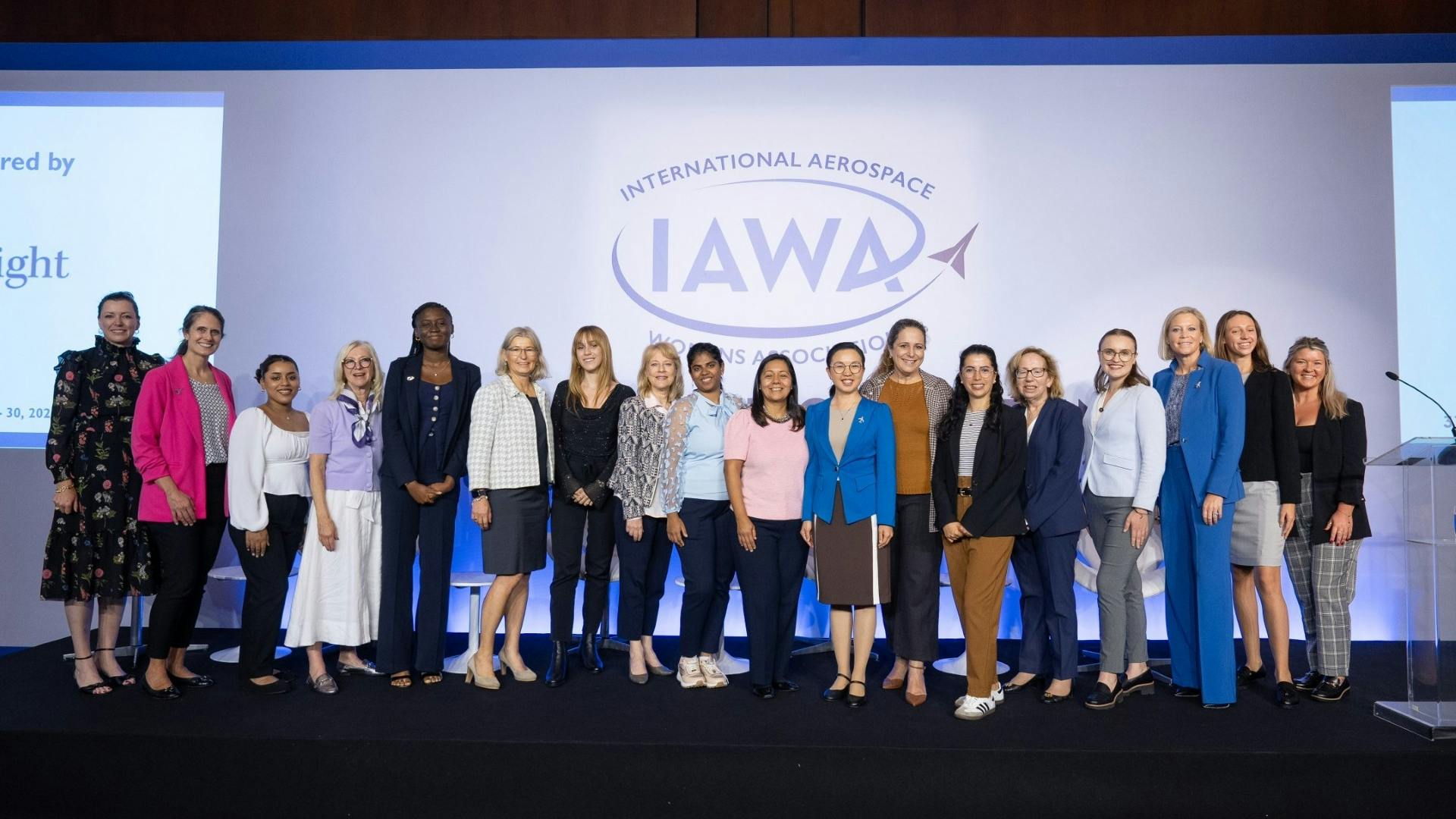 A group of women in business attire stand on a stage smiling at the camera in front of a backdrop that reads, 'International Aerospace Womens Association (IAWA)'