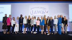 A group of women in business attire stand on a stage smiling at the camera in front of a backdrop that reads, 'International Aerospace Womens Association (IAWA)'