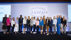 A group of women in business attire stand on a stage smiling at the camera in front of a backdrop that reads, 'International Aerospace Womens Association (IAWA)' A group of women in business attire stand on a stage smiling at the camera in front of a backdrop that reads, 'International Aerospace Womens Association (IAWA)'
