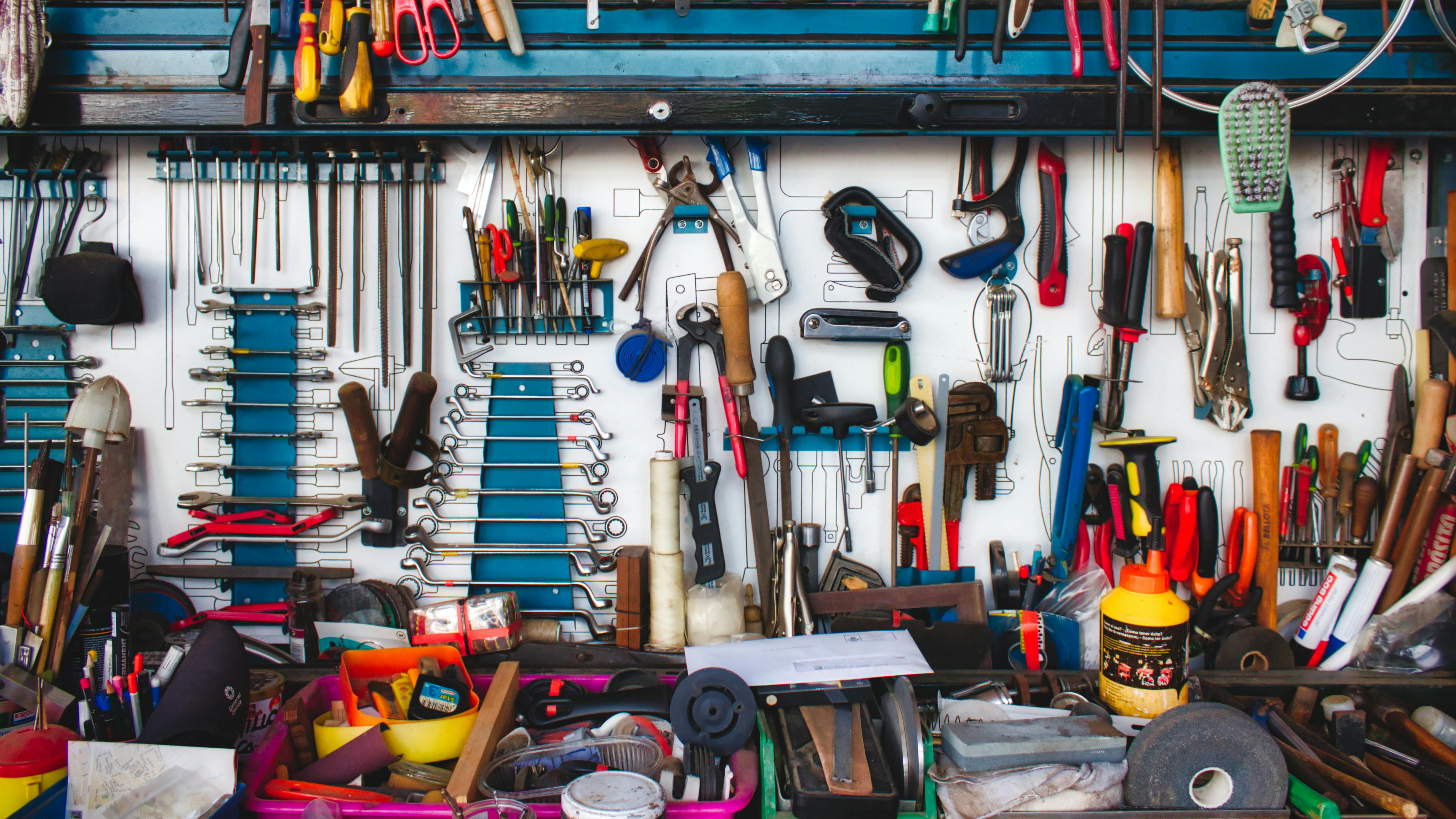 A tool bench with a blue shelf holds a large array of tools, including wrenches, sockets, hammers, pliers and scissors, all arranged messily on top of each other.