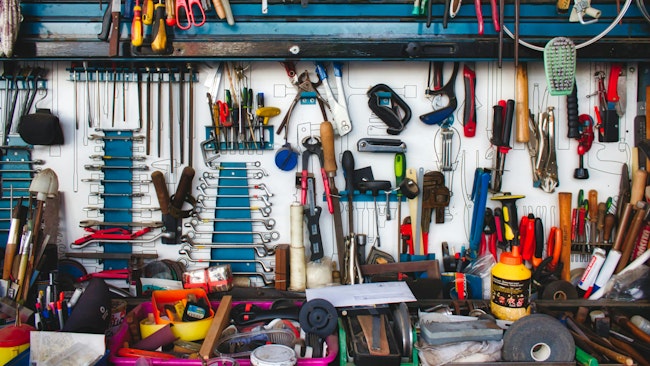 A tool bench with a blue shelf holds a large array of tools, including wrenches, sockets, hammers, pliers and scissors, all arranged messily on top of each other.