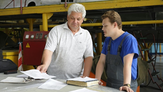 Two aircraft mechanics stand at a table in a repair shop with pieces of paper and a notebook on the table, with one technician speaking and the other holding a piece of paper and the book