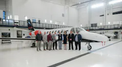 A group of professionals in business attire stand in front of an airplane in an airplane hangar, smiling at the camera