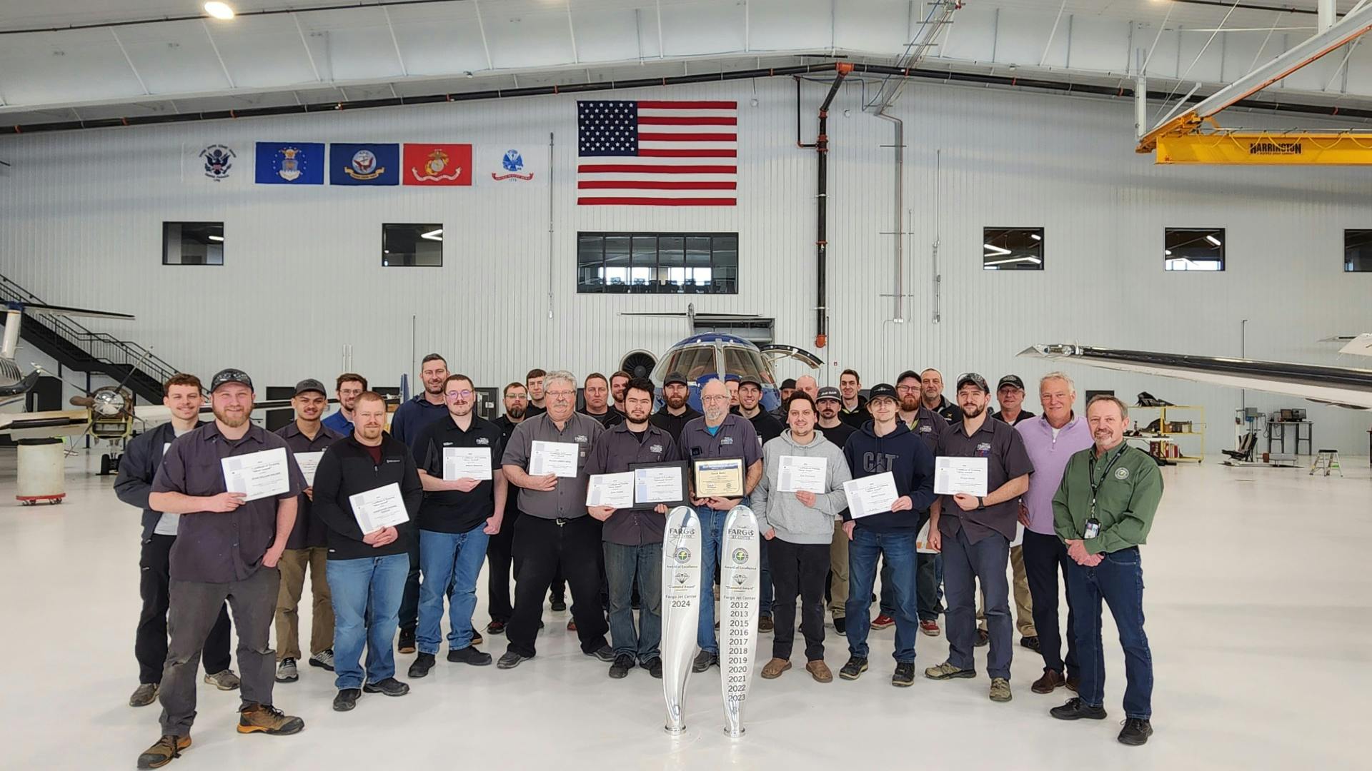 A large group of people stands in an aircraft maintenance hangar in front of three airplanes smiling at the camera with one individual holding a certificate