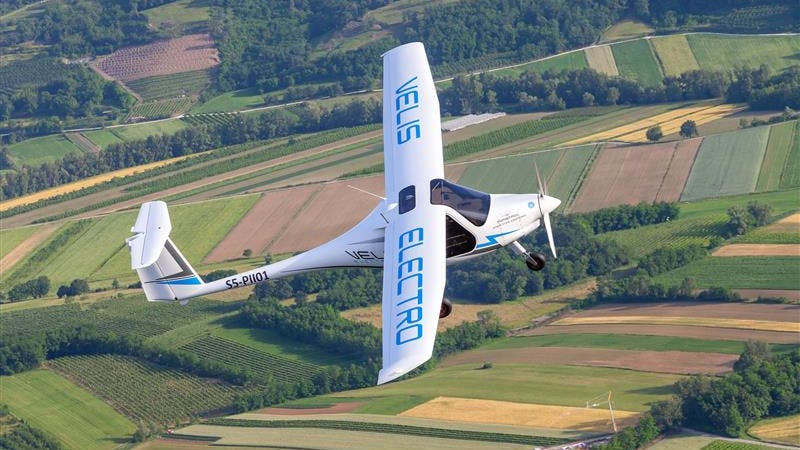 A white airplane featuring blue text that reads, 'VELIS ELECTRO' flies over rows of brown and green fields
