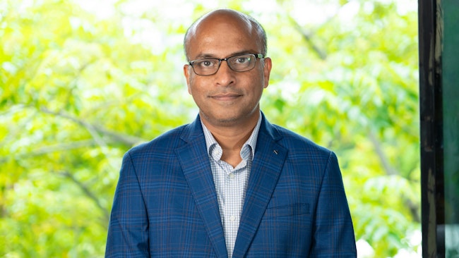 A man wearing a blue and white button-up shirt, glasses, and a blue jacket smiles at the camera