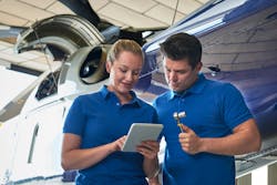 Two aircraft maintenance technicians wearing blue polos stand in front of an airplane looking at a mobile tablet, with one technician holding a socket wrench Two aircraft maintenance technicians wearing blue polos stand in front of an airplane looking at a mobile tablet, with one technician holding a socket wrench