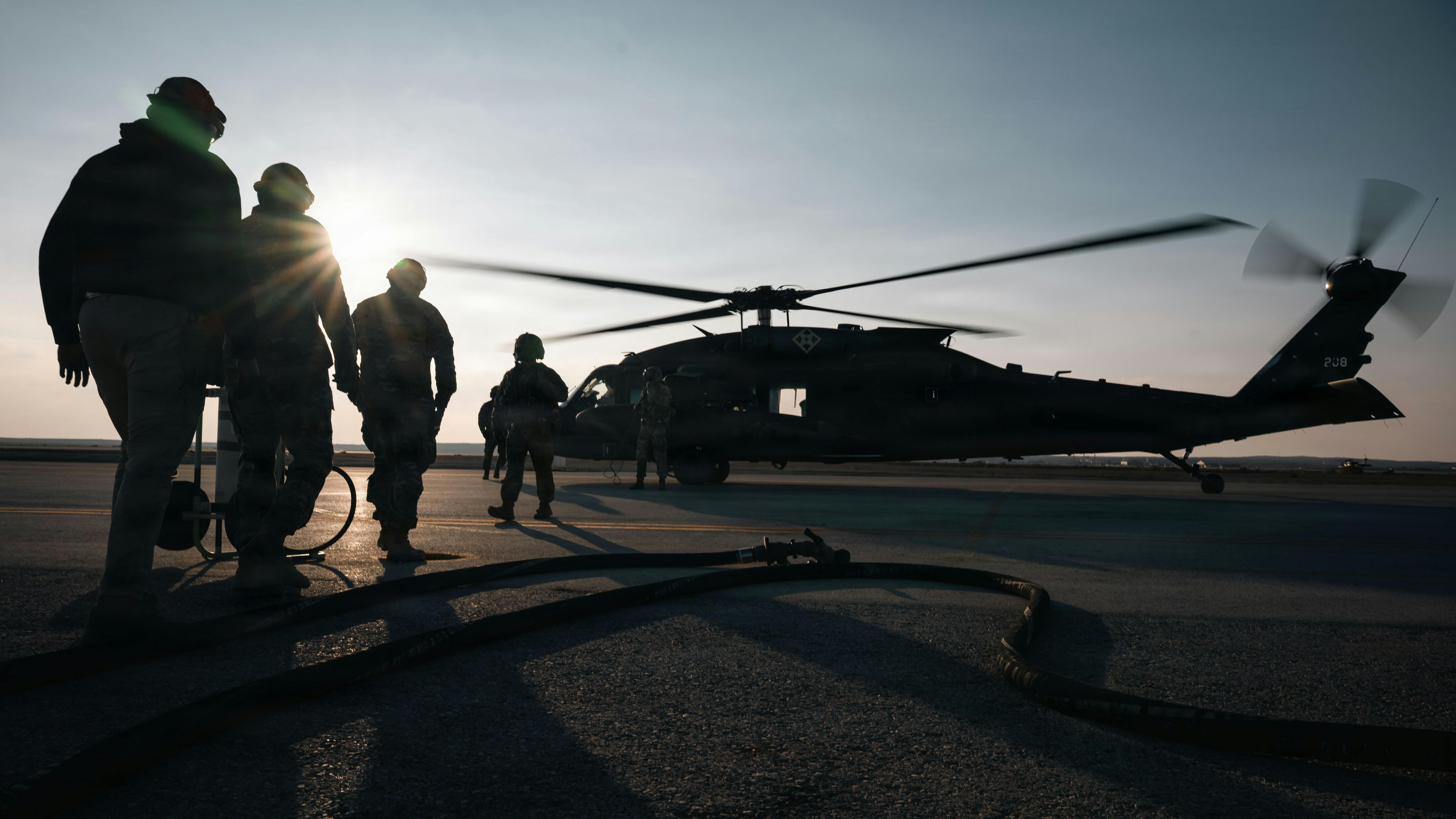 A group of soldiers wearing military dress stand silhouetted near a helicopter that's on the ground with its propellers spinning