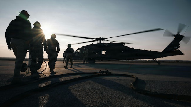 A group of soldiers wearing military dress stand silhouetted near a helicopter that's on the ground with its propellers spinning