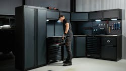 A man in black clothing standing next to a large system of black cabinets and shelves, with a tool box on the top of one of the shelves A man in black clothing standing next to a large system of black cabinets and shelves, with a tool box on the top of one of the shelves