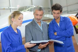 A female AMT in a blue uniform talks with a male AMT in a gray uniform while standing in front of an airplane and reading out of a book together A female AMT in a blue uniform talks with a male AMT in a gray uniform while standing in front of an airplane and reading out of a book together