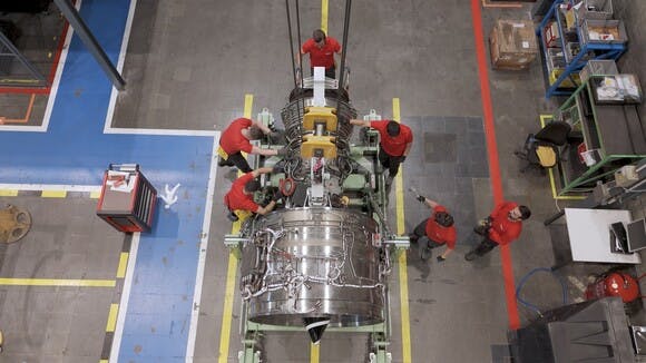 An aerial shot of a group of aircraft mechanics wearing red shirts working on a chrome-colored airplane engine, surrounded by shop equipment