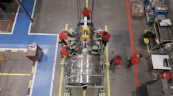An aerial shot of a group of aircraft mechanics wearing red shirts working on a chrome-colored airplane engine, surrounded by shop equipment