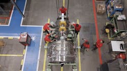 An aerial shot of a group of aircraft mechanics wearing red shirts working on a chrome-colored airplane engine, surrounded by shop equipment An aerial shot of a group of aircraft mechanics wearing red shirts working on a chrome-colored airplane engine, surrounded by shop equipment