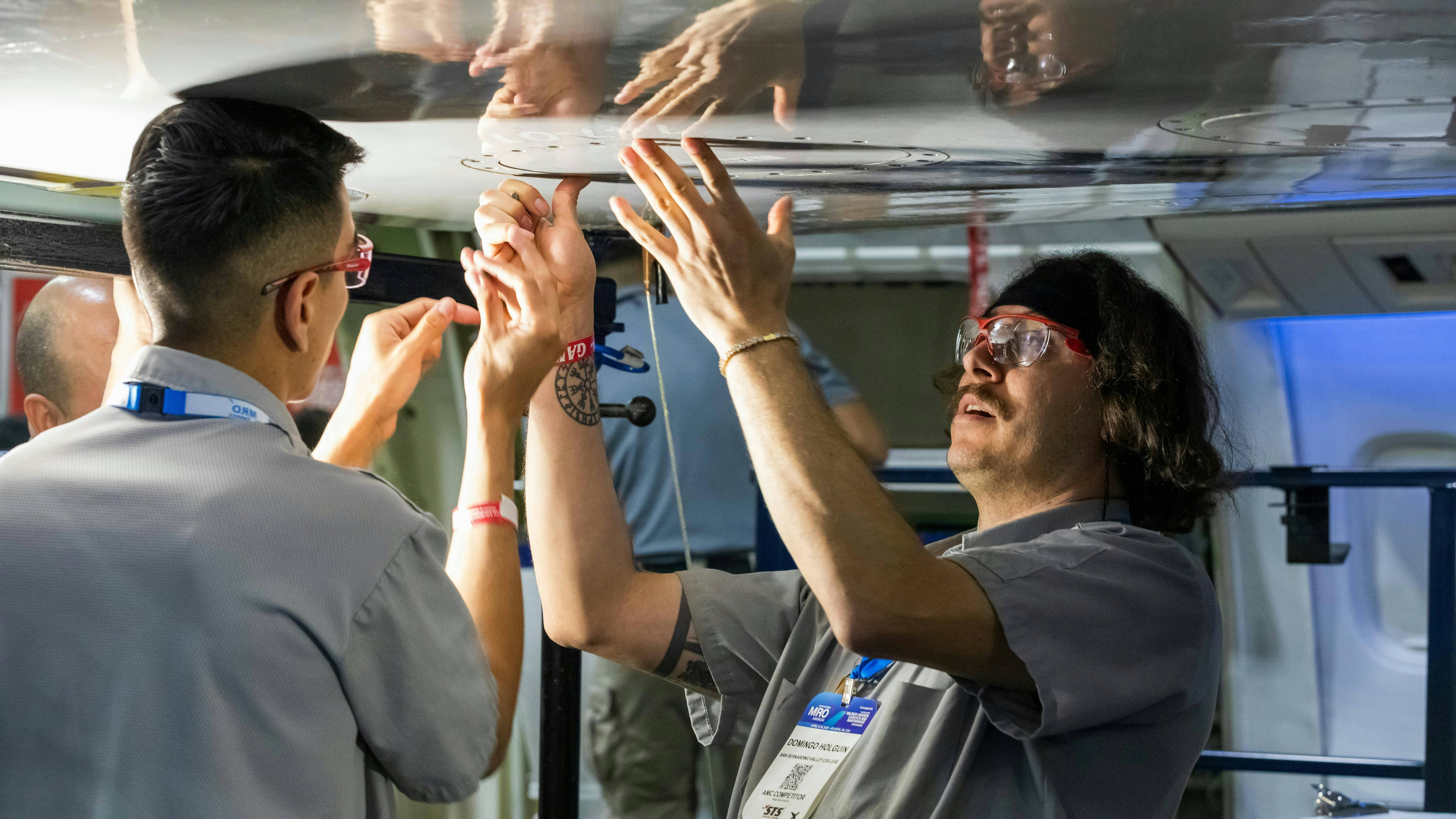 Two aircraft maintenance technicians work on an airplane part while standing under it, a flashlight shining on the part